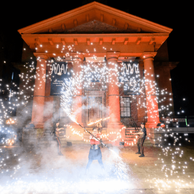 Three people at the Royal Albert Dock, stood in front of a large building with red pillars doing fire dancing.