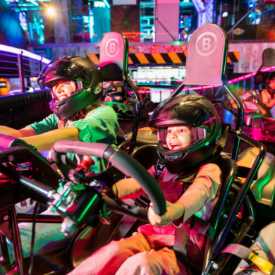 A woman and a boy wearing helmets and sat next to eachother driving a go kart in Gravity Max.