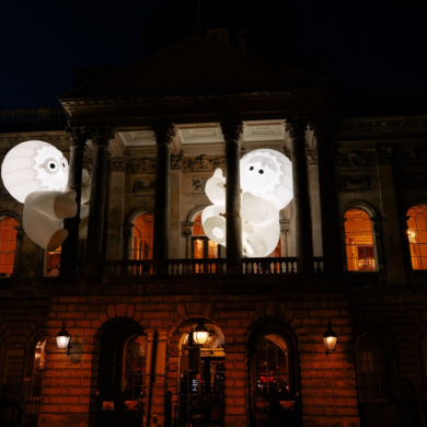 Two large glowing inflatable figures with cartoon-style eyes appear suspended between the columns of a grand historic building at night. The illuminated white sculptures contrast against the dark sky, while warm orange light shines through the building’s windows, creating a playful and dramatic light installation.