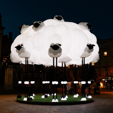 A large illuminated sculpture resembling a flock of sheep forms a glowing white cloud in a city square at night, with individual sheep heads and legs extending from the installation.