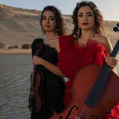 Two sisters stand by a large lake surrounded by sandy hills. The sister on the right is wearing a red dress and holding a cello. The sister on the left wears a black dress and holds a violin.