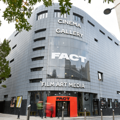 The entrance to FACT with threes lining the side of the building and a red arch leading people into the entrance of FACT.
