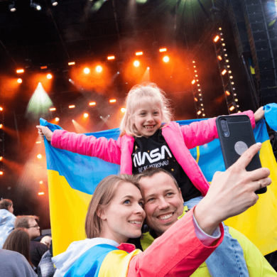 Two adults with a child on their shoulders holding a Ukrainian flag with a large stage behind them.