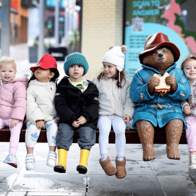 Six young children sitting on a bench with a statue of Paddington.