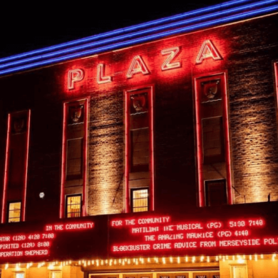The exterior of Plaza Community Cinema. The name of the cinema is lit up red with red writing underneath announcing which shows are on and the times.