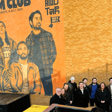 A mural of the band Red Rum Club painted in orange tones on the side of a brick wall.