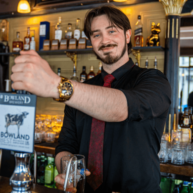 A person stood behind a bar pulling a pint of beer.