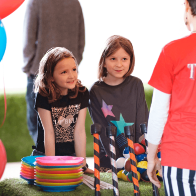 Two children stood at a table with balloons and entertainment going on around them.