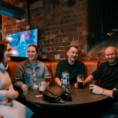 A group of five people sitting round a wooden table with a few drinks on the table. They are smiling and in conversation with a sports match on the tv in the background.