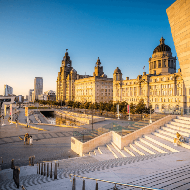 Liverpool Pier Head at golden hour. The sky is very blue and the golden sunlight is reflecting off the Liver Building, Cunard Building and Port of Liverpool. There's a cruise ship docked