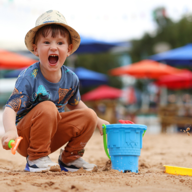 A young child crouches on a sandy play area, holding a small orange spade beside a blue bucket. The child wears a light-coloured hat, a patterned T-shirt, rust-coloured trousers, and trainers. Sand is scattered in mid-air near the bucket, with a yellow toy partially buried nearby. In the background, there are blurred colourful umbrellas and outdoor seating, suggesting a beach-style play zone or family attraction.