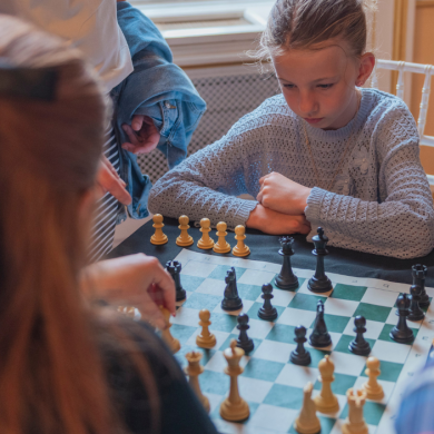A young child sits at a table concentrating on a chess game, with a chessboard and pieces arranged in mid-play. The child rests both arms on the table while looking closely at the board. Several other people stand or sit around the table, partially visible, with one person’s hand reaching toward the pieces as if making a move. The chessboard contains both light and dark pieces scattered across the squares, suggesting an ongoing match. The setting appears to be indoors, with warm lighting, a window in the background, and light-colored walls and chairs. The overall scene shows a small group gathered around the chessboard, focused on the game.