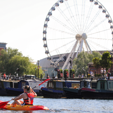 A person paddles a red and yellow kayak across the water at Liverpool’s Royal Albert Dock. Several narrowboats are moored along the waterfront, while the Liverpool Wheel rises prominently in the background. Historic brick dockside buildings, including one displaying signs for “Albert Dock” and “Atlantic Pavilion,” frame the scene on a bright sunny day.