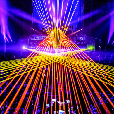 A large indoor concert or performance in a grand hall, viewed from above, with a crowd gathered below. Bright orange, yellow, white, and purple laser beams cross dramatically over the audience, forming geometric patterns that converge toward a central stage. Purple stage lighting illuminates the ornate architecture on both sides of the venue, creating a vibrant, high-energy atmosphere.