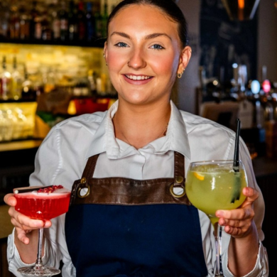 Person standing behind a bar counter holding two colourful cocktails, one red and one pale green, in stemmed glasses. The person is wearing a light shirt and dark apron, with shelves of bottles, glassware and illuminated bar displays visible in the softly blurred background.