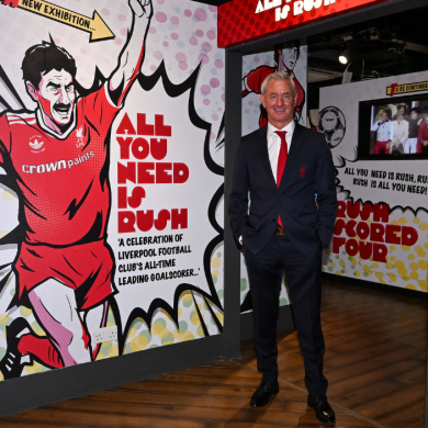 A Ian Rush stands inside a Liverpool Football Club museum exhibition beside a large graphic display celebrating the club's all-time leading goalscorer. The exhibition features bold red-and-white comic-style artwork, football memorabilia, trophy displays and interactive screens, with prominent text reading “All You Need Is Rush.”