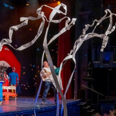 A lively stage show inside an auditorium where performers launch long streams of white paper into the air, creating swirling ribbons above the stage. Confetti falls as one performer stands holding equipment that appears to fire the paper, while another stands nearby with a guitar prop. The set includes colourful backdrops, including a large red lips display and a science-themed sign, with audience seating visible to the right under warm stage lighting.