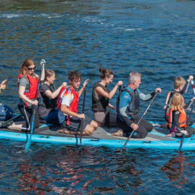 Eight people wearing life jackets kneel on a large blue paddleboard and use paddles to move across calm water. The group includes adults and children, and the board is viewed from the side with reflections visible on the water's surface.