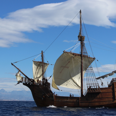 A large wooden sailing ship with multiple masts and cream-colored sails travels across a calm blue sea under a bright sky. Mountains are visible in the distance along the horizon, and soft white clouds stretch across the background. The ship is viewed from the side, showcasing its historic design, dark timber hull, and intricate rigging.