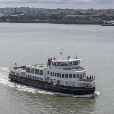 A white Mersey Ferry travels across the River Mersey, creating a trail of wake behind it. The vessel is viewed from above and to the side, with the opposite riverbank visible in the background, featuring waterfront buildings, trees and a ferry terminal. The water is calm under an overcast sky, with soft sunlight reflecting on the river’s surface.