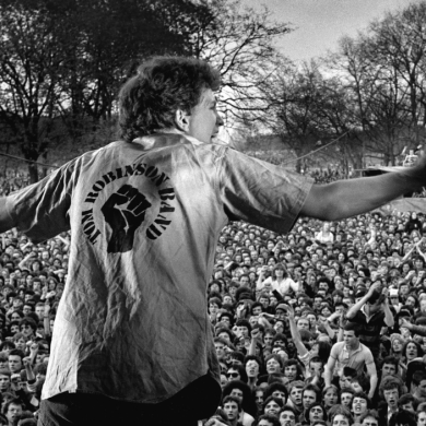 Black-and-white photograph of a performer standing on an outdoor stage with arms outstretched, facing a large crowd gathered in a park. The back of the performer's shirt features a raised fist graphic and the words “John Robb’s on Band.” Thousands of audience members fill the foreground and background, with leafless trees visible behind the crowd, creating the atmosphere of a major live music event.