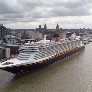 A large cruise ship sails along Liverpool’s waterfront on the River Mersey, with the city skyline and historic waterfront buildings visible in the background beneath a dramatic cloudy sky.