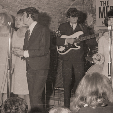 Vintage black-and-white photograph of a live music performance in a cellar-style venue. Several performers stand and sing at microphones while a musician plays guitar in the background. Audience members are visible in the foreground, and a sign for “The Fabulous Merseybeats” hangs on the wall behind the stage.