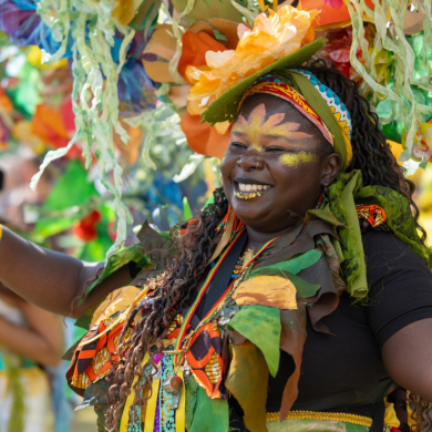 Performer in a vibrant carnival costume decorated with large colourful flowers, leaves and streamers, raising one hand while taking part in a parade. The costume features tropical-inspired green and orange elements, face paint and floral headpieces. Brightly coloured decorations fill the background, creating a lively festival atmosphere.