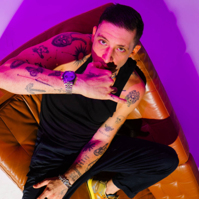 A studio portrait of a person sitting diagonally on a tan leather armchair, viewed from a slightly elevated angle. The person is wearing a black sleeveless top, black trousers, and yellow trainers. One arm rests across the lap while the other is raised, with the hand positioned near the mouth in a gesture resembling a phone call sign. The person has multiple visible tattoos on both arms, including various small designs and symbols. A wristwatch with a metallic strap is worn on one wrist, along with a bracelet.
The background is minimal and clean, with strong coloured lighting creating a gradient effect—magenta/pink tones dominate one side while a softer purple and white blend appears on the other. The lighting casts bold shadows behind the chair, adding contrast and depth to the composition.
The leather chair has a soft, slightly wrinkled texture, and its warm brown colour contrasts with the cool-toned lighting. No text is visible in the image.