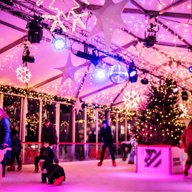People ice skating inside a festive indoor rink decorated with twinkling fairy lights, illuminated snowflake ornaments and a large Christmas tree. The rink is bathed in pink and purple lighting, creating a vibrant winter atmosphere.
