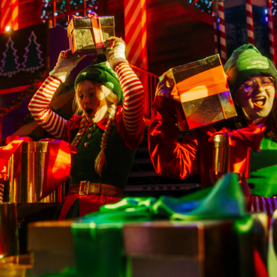 Festive performers dressed in green elf costumes and red-and-white striped sleeves hold wrapped presents inside a brightly lit Christmas-themed venue. Colourful decorations, wrapped gifts and glowing red and green lights create a lively holiday scene.