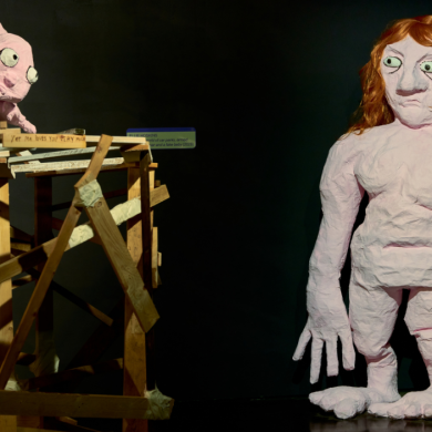 Museum installation featuring two large handmade papier-mâché figures in a dark gallery space. One human-like figure with red hair stands in the foreground, while a pink rabbit-like creature crouches on a wooden scaffold structure to the left. The exhibit is illuminated by spotlights, highlighting the rough textures and sculptural details against a black backdrop.