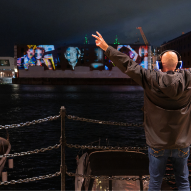 Person wearing illuminated headphones stands on Liverpool’s waterfront at night, facing a large building displaying colourful projected artwork across its facade. Boats are moored in the foreground, with reflections shimmering on the water beneath the city lights.