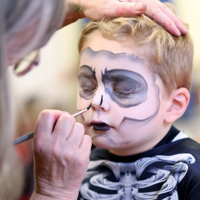 A child sits still while face paint is carefully applied to create a skull design. The close-up image shows detailed black and white face paint around the eyes, nose and mouth, with an adult’s hand steadying the child’s head. The background is softly blurred, focusing attention on the face-painting activity.