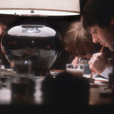 Warmly lit indoor scene showing three people seated around a table under a large table lamp, concentrating on painting or detailed craft work. Each person holds a thin paintbrush and works over small items or surfaces in front of them. The lamp’s soft light reflects off a round, dark lamp base at the centre, while the surrounding table holds cups, tools, and materials. The background is dim, drawing attention to the focused activity and hands-on detail at the tabletop.