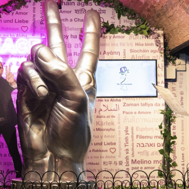 Large metallic sculpture of a hand making a peace sign displayed indoors beneath a brick arch, surrounded by decorative greenery and a patterned wall featuring the word “peace” written in multiple languages. A life-size cutout figure stands to the side, also holding up a peace sign. Two screens and informational plaques are positioned around the display, with pink and purple lighting illuminating the scene.