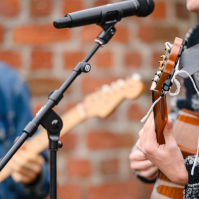 Close-up of musicians performing live, with an acoustic guitar in the foreground and an electric guitar slightly out of focus behind, both playing into a microphone against a brick wall backdrop.