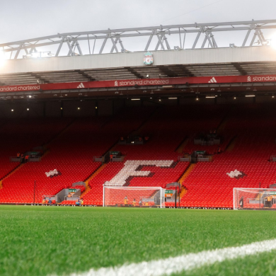 Wide view of Anfield stadium with empty red seating spelling “L.F.C.” in white. Bright stadium lights shine along the roofline, while the green pitch and a white sideline are visible in the foreground. Goals, equipment and a few staff are seen near the stands.