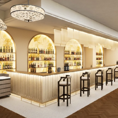 Interior view of a modern bar and dining space with a long wooden bar counter and high stools. Shelving behind the bar displays rows of bottles set into arched, backlit alcoves. Warm neutral colours, patterned light fittings, and polished wooden flooring create an elegant atmosphere. Tables and chairs are arranged to the right side of the room.