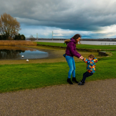 An image of a woman holding a childs hand and playing by swinging them round. They are standing on a path with a large lake behind them.