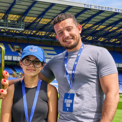 A man and a young girl are standing smiling and looking at the camera. The football stadium is behind them, with the young girl wearing a blue hat, and both have blue lanyards around their neck.