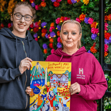 Two people holding a poster for Southport Beer Week infront of a wall covered in flowers.
