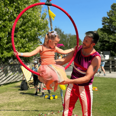 A child sitting in a large hoop used in acrobatics whilst and adult wearing bright pink clothing holds the hoop.