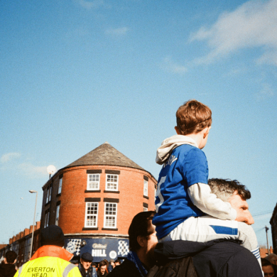 A photo of a young boy sitting on an adult's shoulders wearing an Everton kit. There is a blue sky in the background and the tops of some buildings.