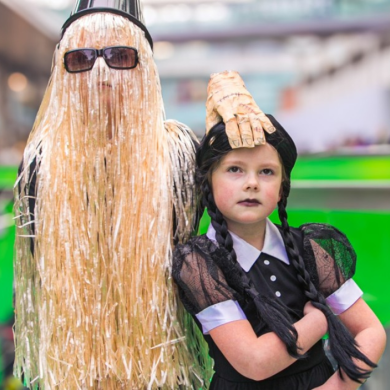 A young girl dressed as Wednesday Addams from the Addams Family with somone dressed up as thing stood beside her.