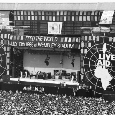 A black and white photo of a concert with a stage set up that says 'Live Aid' on either side of the stage. A large crowd is in front of it.