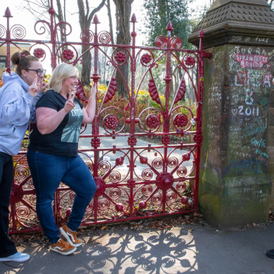 Two women smiling and posing while having their picture took by a man in-front of the Strawberry Field gates.
