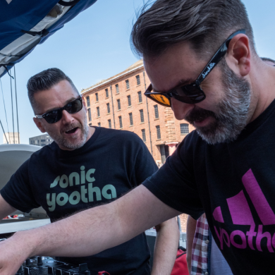 2 men are DJing. They both wear sunglasses and black t-shirts with writing. One of their shirts says 'Sonic Yootha''