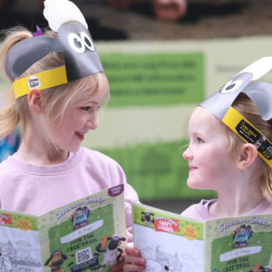Two young children wearing paper headbands themed around Shaun the Sheep stand facing each other while holding open activity maps or brochures. The children are dressed in light pink tops and are positioned in front of a softly blurred indoor background with green signage. The printed maps include illustrations, text, and a visible QR code related to a trail or activity.