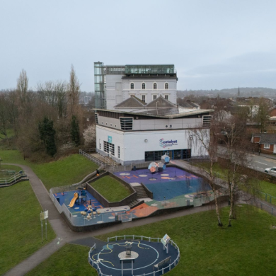 Aerial view of a modern white building with the sign “Catalyst” on the front, set beside a colourful playground with rubber flooring, slides, and play structures. The site is surrounded by grassy parkland with walking paths and a small circular play area in the foreground. To the left, a river runs alongside trees and open green space, while to the right there are residential houses and a road with parked cars. In the background, a large green arch bridge rises over the landscape under an overcast sky.